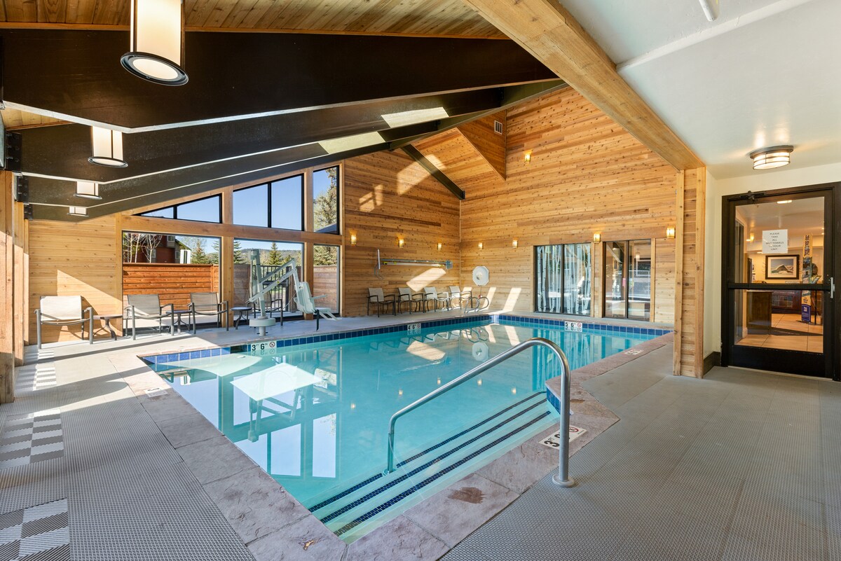 An indoor swimming pool is featured, surrounded by large windows allowing natural light to illuminate the wooden walls. Comfortable lounge chairs are positioned nearby for relaxation, and a sliding glass door leads to an outdoor area.