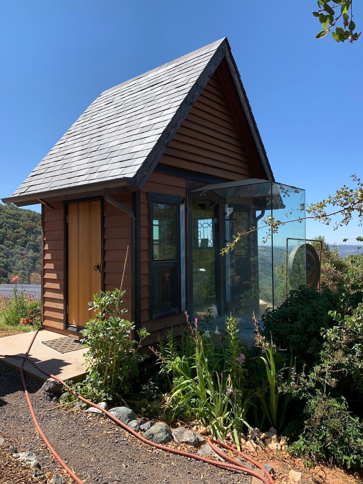 A charming structure is featured with wooden siding and a sloped roof, complemented by large glass panels that enhance the view of the surrounding landscape. Lush greenery is visible around the entrance, creating a seamless connection with nature.