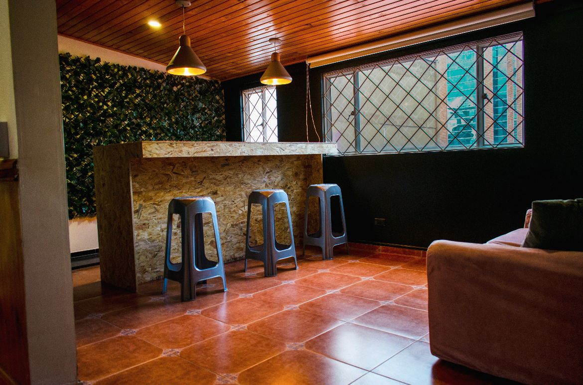 A modern bar area features a textured stone counter with three metal stools. Greenery adorns the wall behind the bar, while a large window allows natural light to brighten the space. The floor is tiled with warm, reddish-brown ceramic tiles.