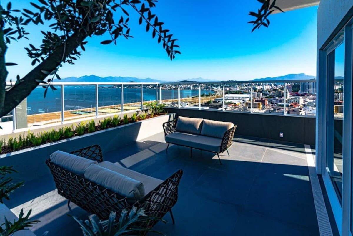 A rooftop terrace is showcased, featuring a seating area with two woven chairs and a cushioned sofa. A panoramic view of the city and surrounding mountains is visible in the background, with bright blue skies enhancing the outdoor space.
