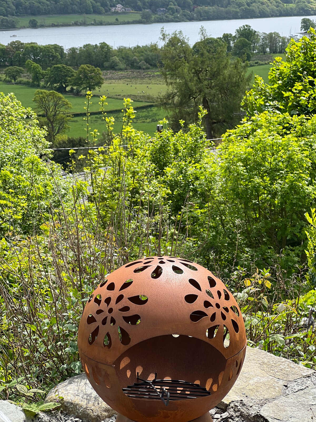 A decorative fire pit is positioned in the foreground, featuring intricate cut-out patterns. Lush greenery surrounds the area, with vibrant foliage framing a scenic view of a lake and rolling hills in the background, inviting relaxation in a tranquil outdoor space.