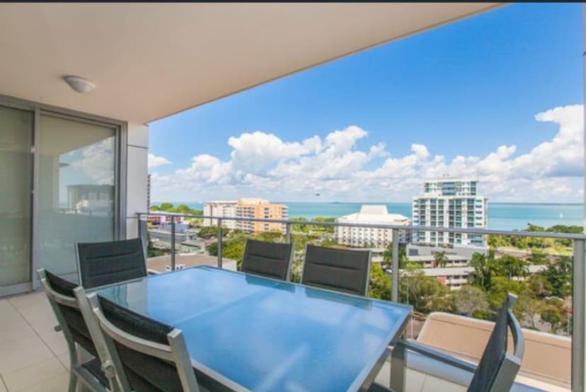 A spacious outdoor balcony features a glass table surrounded by six chairs, offering an unobstructed view of the harbor and surrounding buildings. The sky is partly cloudy, with hints of blue peeking through, creating a bright and open atmosphere.