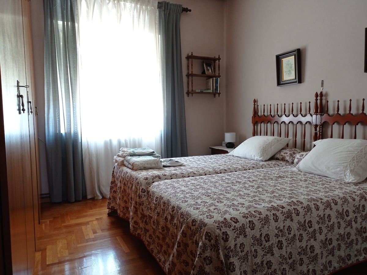 A well-lit bedroom features two twin beds dressed in patterned linens. A bright window is framed by sheer curtains, allowing natural light to fill the room. Decorative elements include a wooden shelf and a framed picture on the wall, providing a cozy touch.