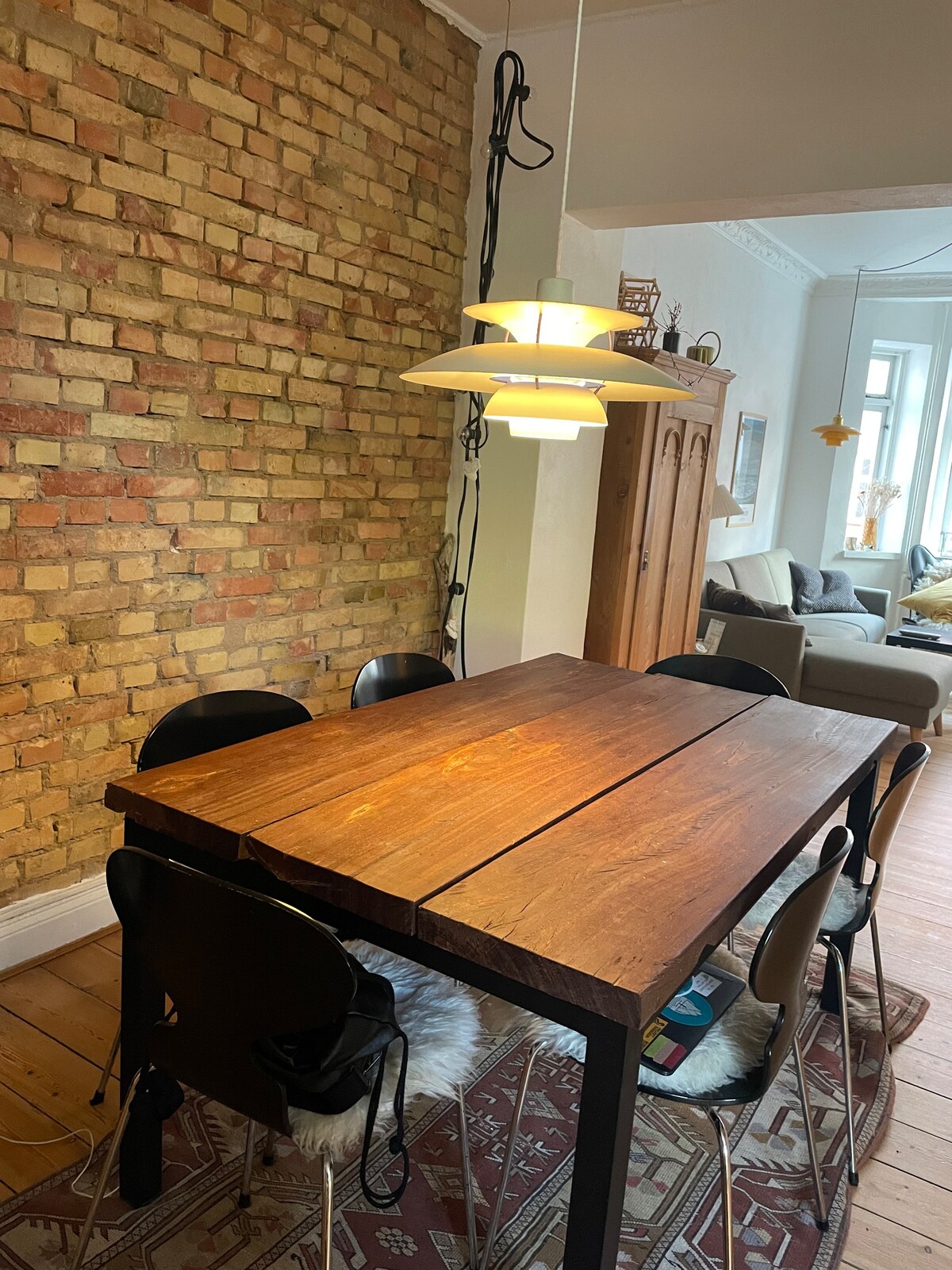 A modern wooden dining table is centrally positioned, surrounded by six black chairs. A unique pendant light hangs above the table, while a textured brick wall provides an appealing backdrop. The space features a glimpse of a comfortable living area visible in the background.