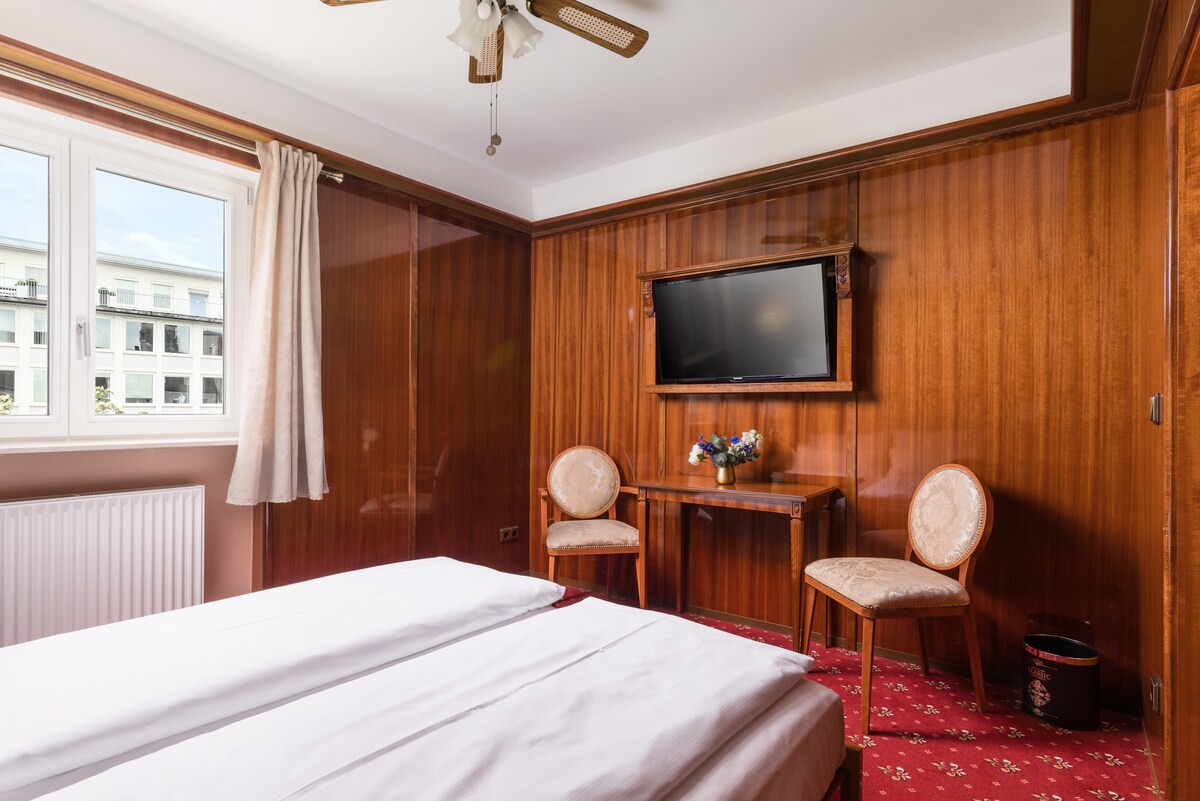 A warm wood-paneled room features a spacious bed with white linens. Two elegant chairs are positioned beside a small table, and a 40" flat-screen TV is mounted on the wall. A large window invites natural light, complemented by light curtains.