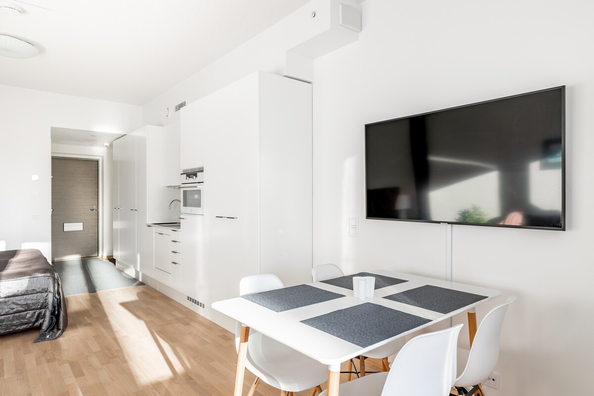 A modern living space features a spacious dining table with white chairs, positioned beside a large television mounted on the wall. The kitchen area is partially visible, showcasing sleek cabinetry. Natural light fills the room, enhancing its bright and uncluttered appearance.