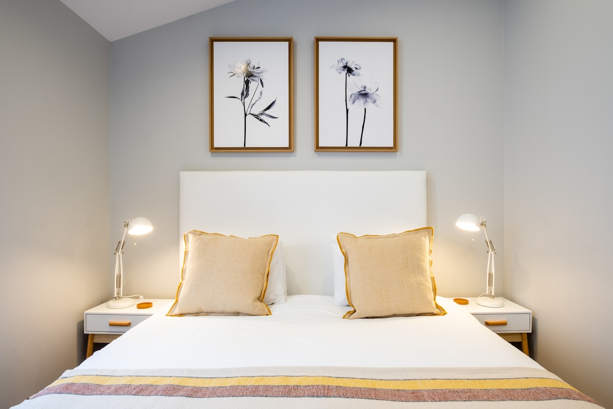A cozy sleeping area features a spacious bed with two decorative pillows and a soft blanket. Light fixtures on either side provide illumination. Two framed floral artworks are displayed above the headboard, adding a touch of elegance to the serene, neutral color palette.
