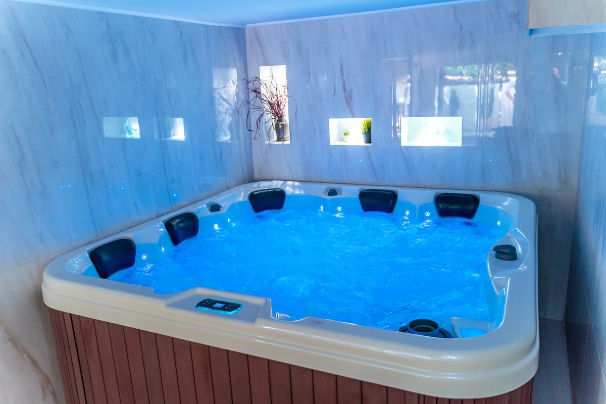 A spacious hot tub is positioned in a well-lit area, surrounded by sleek marble walls. The blue water invites relaxation, while dim blue lighting enhances the tranquil environment. Small shelves display minimalist decor, contributing to a serene ambiance.