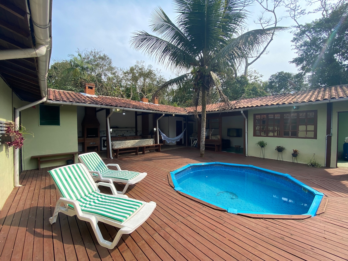 An inviting outdoor area features a private pool surrounded by a wooden deck, complemented by lounge chairs with striped fabric. Palm trees provide shade, while the nearby entrance leads to an open living space, creating a relaxing atmosphere for leisure.