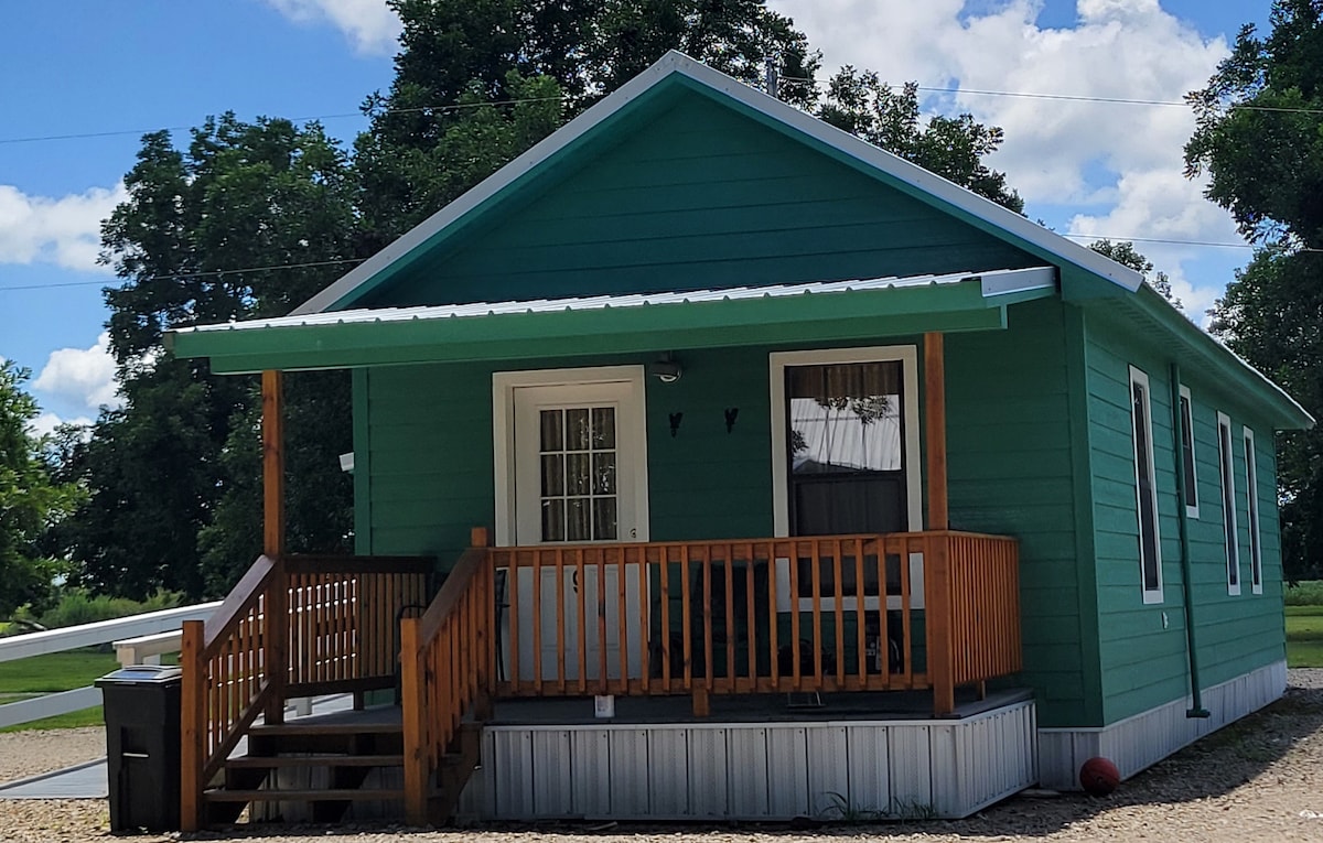 A charming green cottage features a wooden front porch with steps leading to the entrance. Large windows invite natural light, and the surrounding greenery enhances the peaceful setting. The structure is situated on a gravel pathway, providing easy access from the outdoor space.