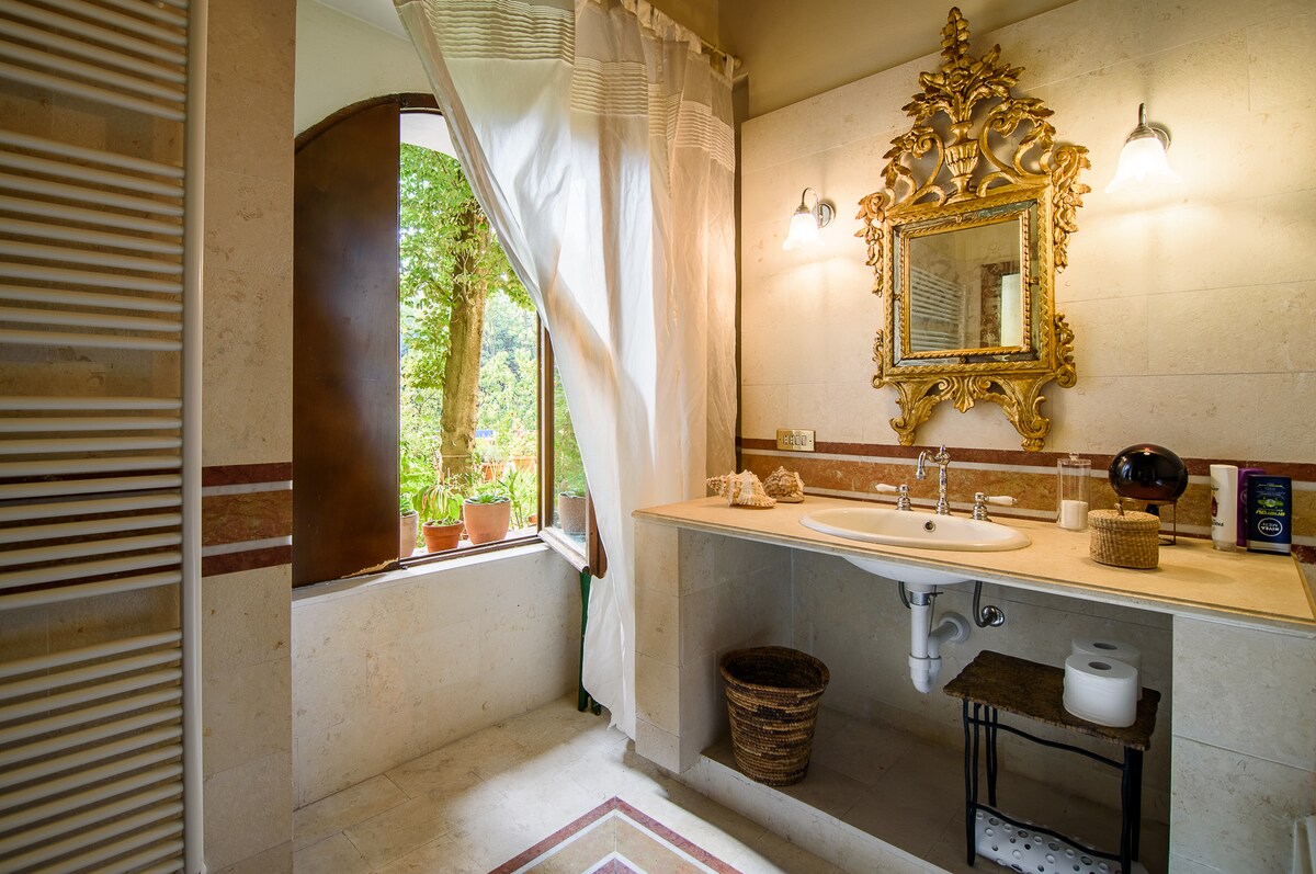 A serene bathroom features elegant stone walls and a large mirror adorned with intricate gold detailing. Natural light filters through a window framed by soft drapes, highlighting potted plants outside. A wooden shelf holds essential toiletries, while a woven basket adds a touch of warmth.