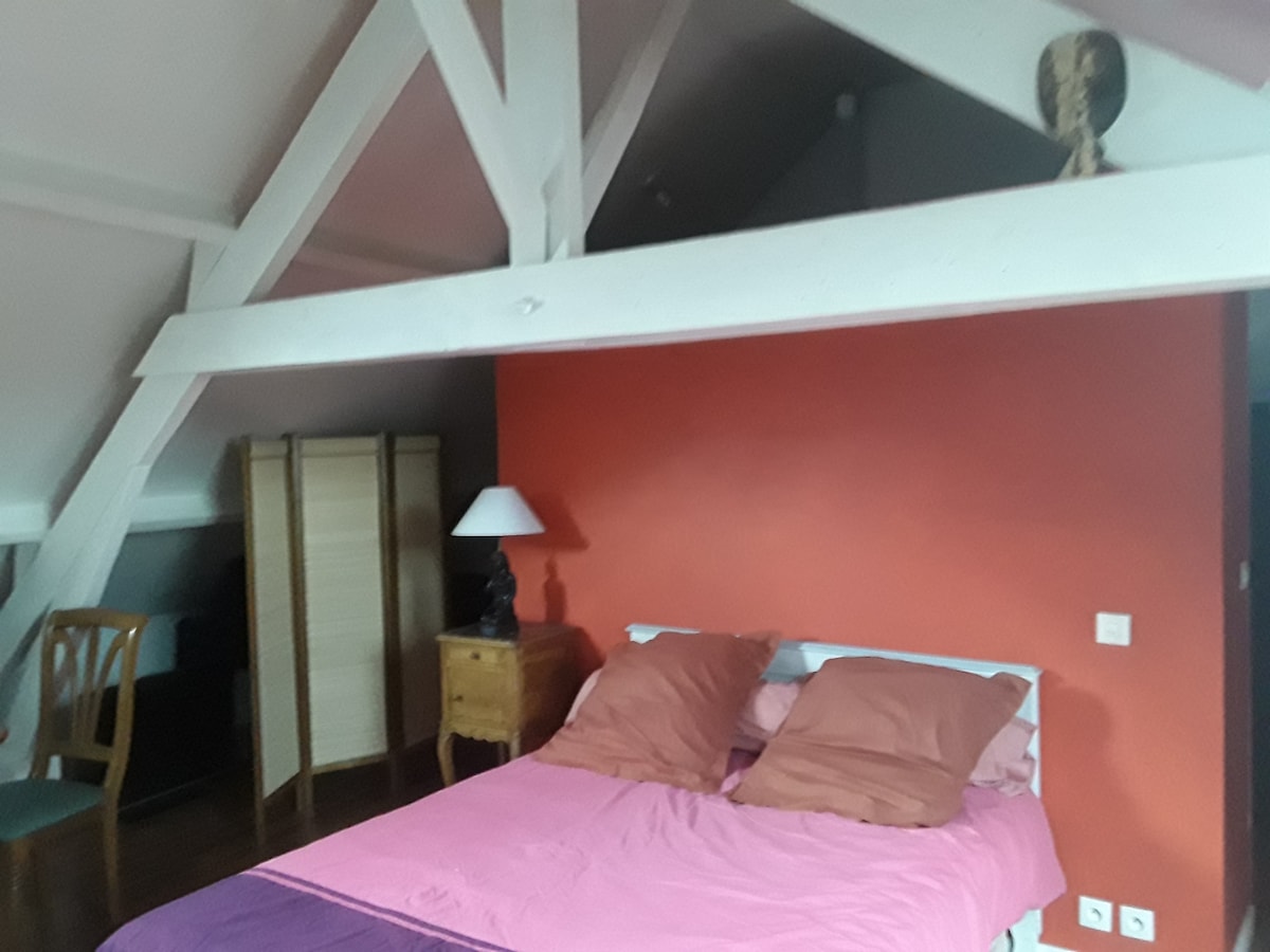 The spacious bedroom features a cozy bed dressed in soft linens. A softly lit lamp sits on a bedside table next to the bed. The room is complemented by a rich orange wall and visible wooden beams that enhance the lofted ceiling.