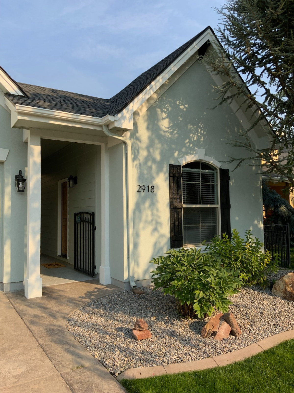The exterior entryway of a guest house is shown, characterized by a light-colored facade and a welcoming pathway. Lush greenery and decorative stones line the walkway, enhancing the outdoor space. A security gate can be seen leading to the entrance, providing a sense of privacy.
