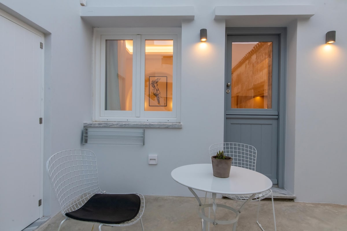 A small outdoor seating area features a round white table complemented by two wire chairs, one with a cushion. Soft lighting from wall sconces illuminates the light grey walls, creating a welcoming entrance. A potted plant adds a touch of greenery to the space.