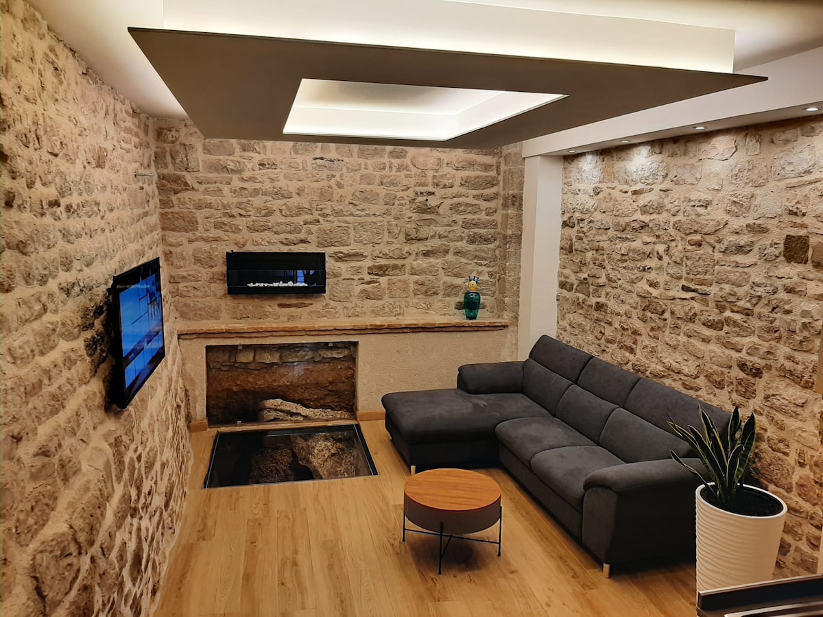 A cozy living area features a grey sectional sofa positioned around a round wooden coffee table. Stone walls display rustic charm, while modern lighting provides a soft glow. A wall-mounted television and a decorative fireplace add functionality and warmth to the space.