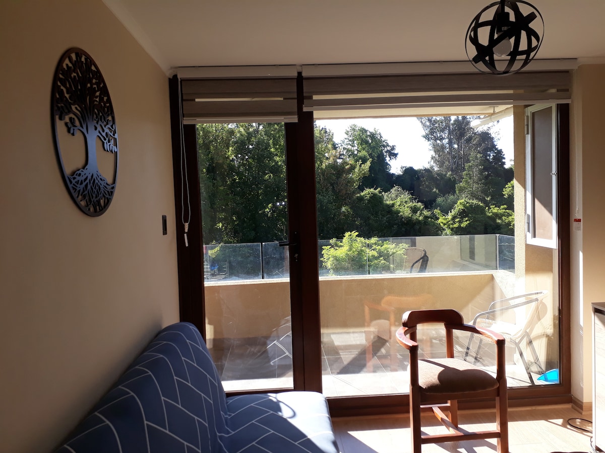 A comfortable seating area is present, featuring a blue patterned sofa and a wooden chair. Large glass doors illuminate the space, opening to a balcony overlooking greenery. The wall displays a decorative piece shaped like a tree, adding a unique touch.