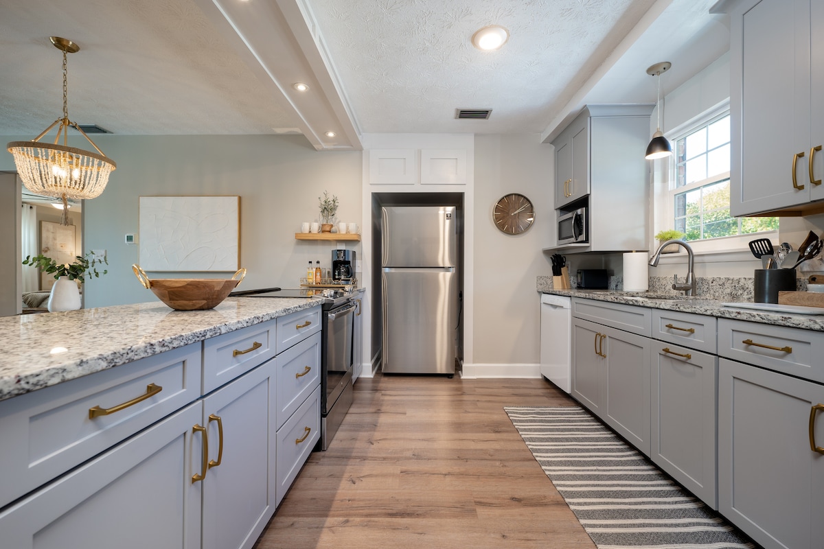 The kitchen features modern cabinetry in soft gray tones, complemented by a large granite countertop. Stainless steel appliances include a refrigerator and microwave. A window allows natural light to brighten the space, while a striped rug adds texture to the floor.