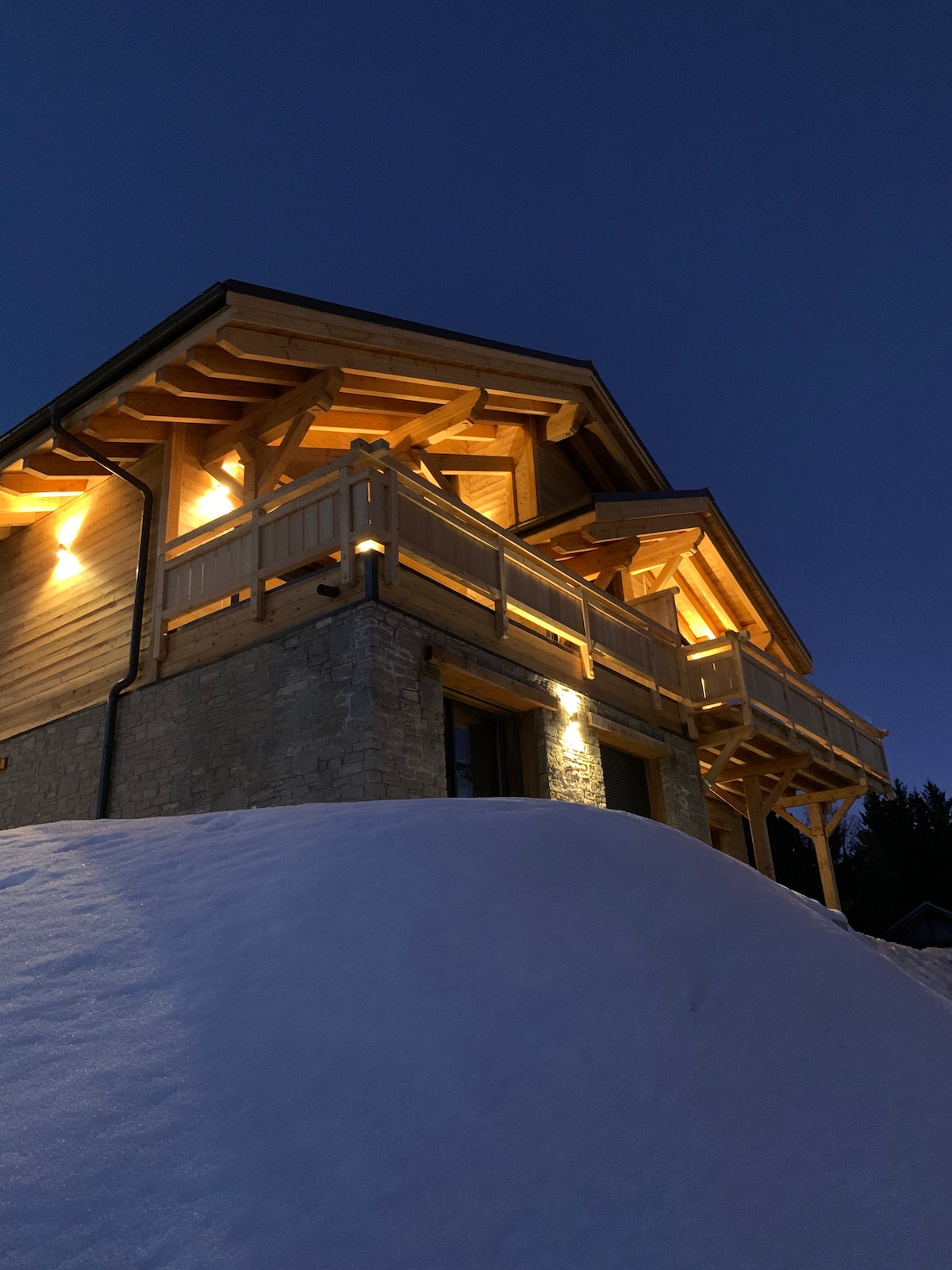 The chalet structure is illuminated at dusk, showcasing a blend of wood and stone materials. A spacious balcony with wooden railings is featured prominently, while a snowy landscape surrounds the base, creating a serene ambiance.