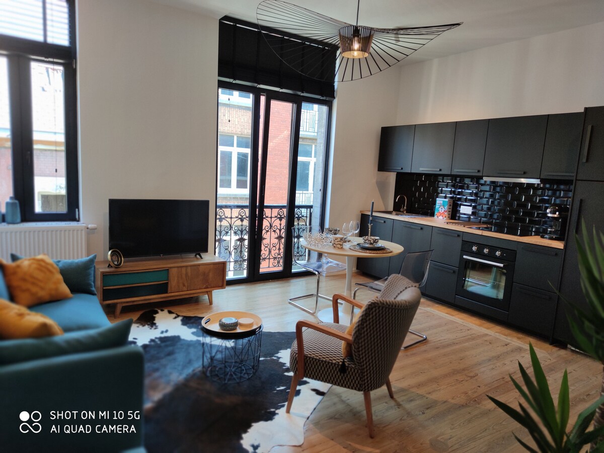 A modern living space features a comfortable blue sofa beside a stylish coffee table. A round dining table with seating complements the sleek kitchen area, equipped with black cabinetry and stainless-steel appliances. Natural light streams through large windows adorned with elegant black frames.