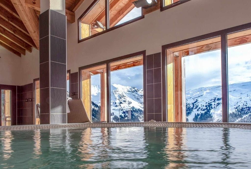 The wellness area features a shimmering spa pool with a view of snow-capped mountains through expansive windows. The natural light accentuates the wooden beams above, creating a serene environment for relaxation.