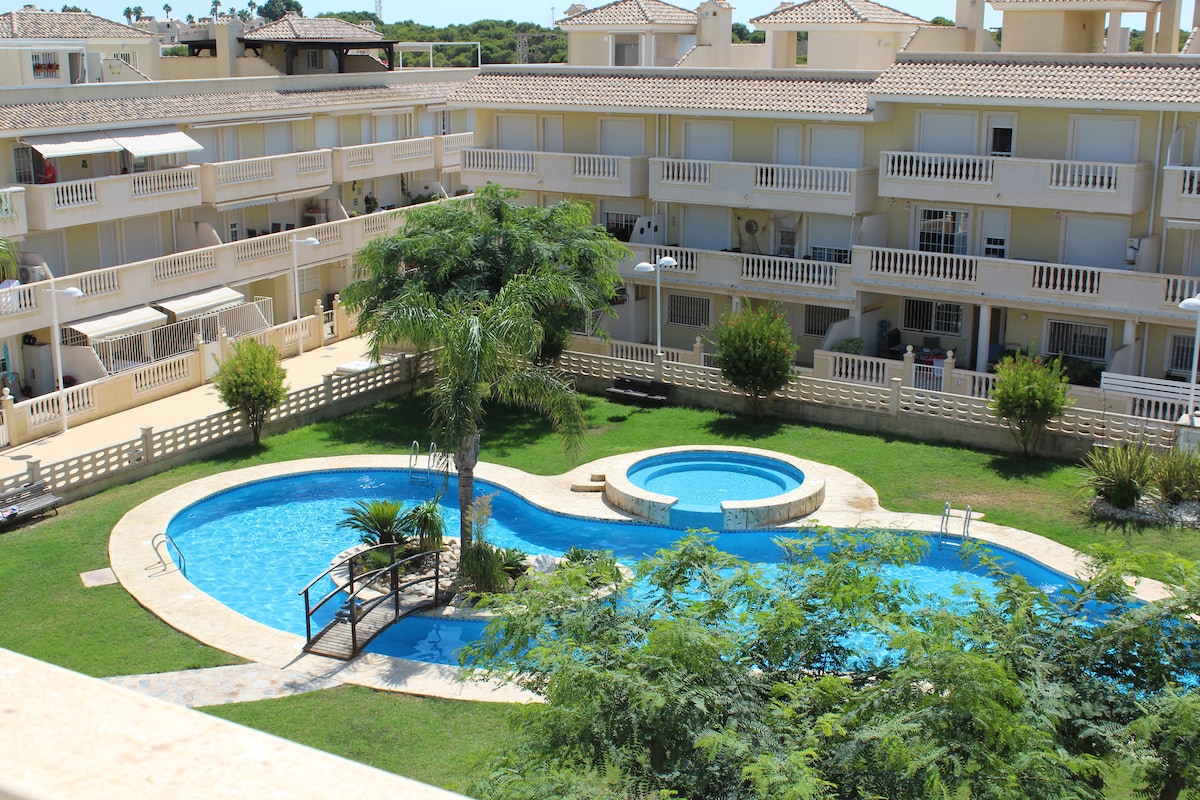 A spacious outdoor area features a large swimming pool surrounded by green lawns and palm trees. A smaller jacuzzi is situated adjacent to the pool, and pathways lead through the landscaped grounds, providing easy access to the pool area.