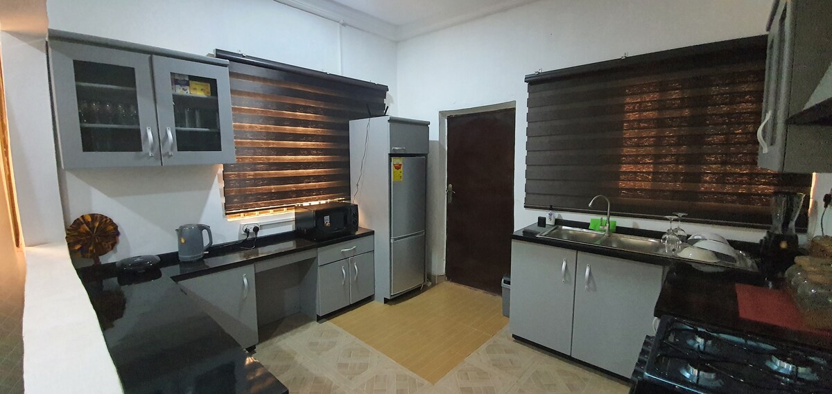 The kitchen features sleek gray cabinets and a black countertop. A refrigerator and a microwave are visible, along with a sink under a window adorned with dark blinds. Natural light filters softly into the space, creating a functional and inviting atmosphere.