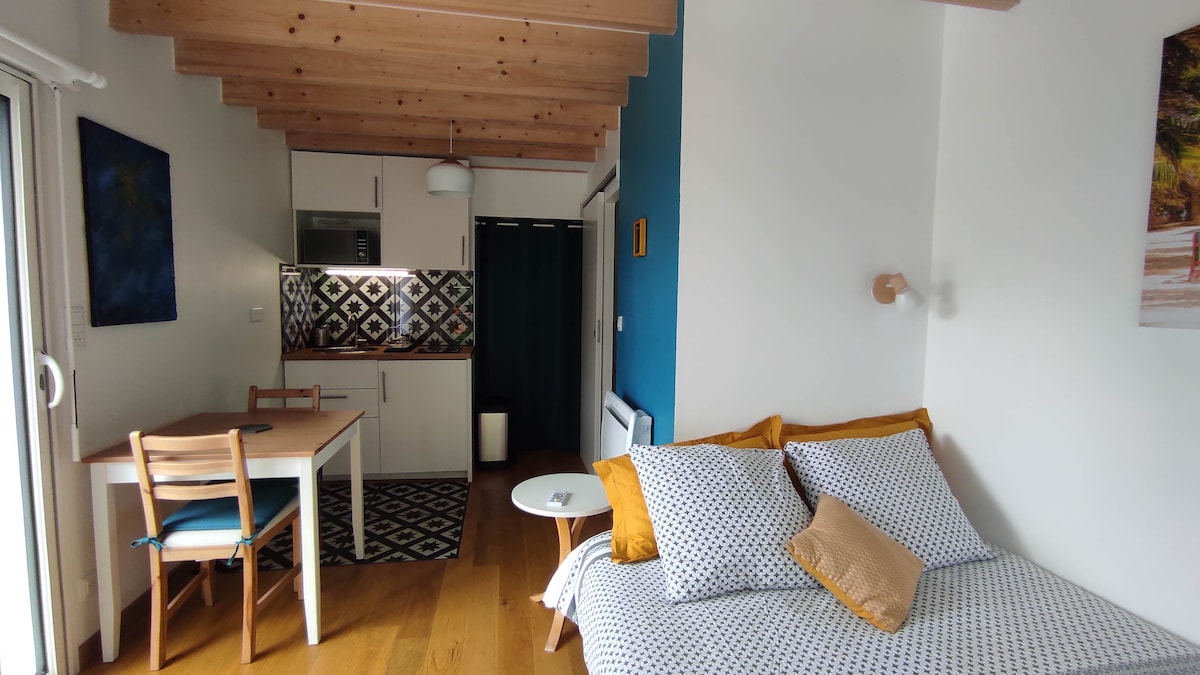 A well-lit studio with wooden beams features a cozy convertible sofa and a dining table. A patterned backsplash complements the kitchen area, while bright blue and neutral tones create a harmonious atmosphere. Natural light floods the space through large windows.