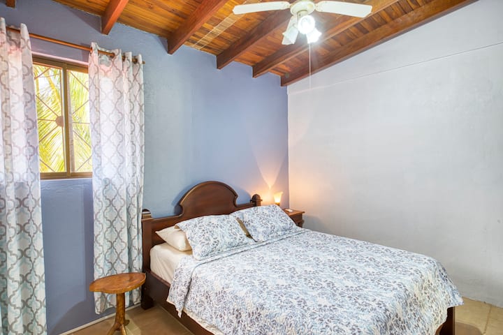 ห้องพักสว่างพร้อมเตียงคู่ที่สะดวกสบาย เพดานไม้อุ่นๆ พัดลมและเครื่องปรับอากาศ การตกแต่งที่เรียบง่ายและผ่อนคลาย เหมาะสำหรับค่ำคืนที่สงบ หน้าต่างพร้อมวิวเขตร้อนและม่านมีสไตล์ 