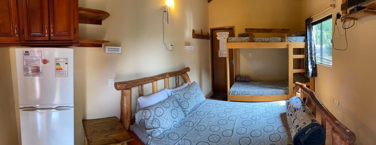 A cozy cabin interior features a spacious bed with a patterned cover and a wooden headboard. A bunk bed is situated in the corner, offering additional sleeping arrangements. Natural light filters in through a window, highlighting the warm wooden accents and simple furnishings.