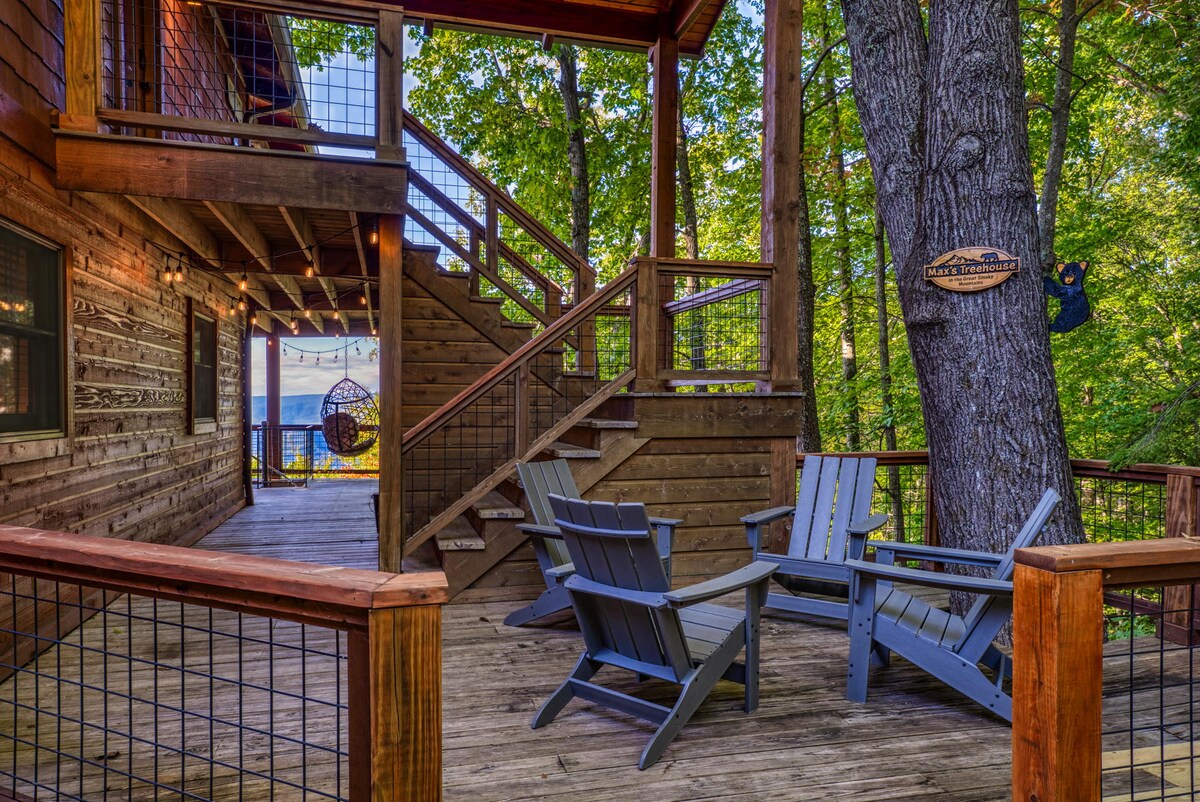 A wooden deck showcases two Adirondack chairs positioned around a natural wood surface. Sturdy stairs lead to an upper level, surrounded by dense trees. A wooden sign affixed to a nearby tree indicates the cabin's name, enhancing the welcoming atmosphere of this forest retreat.