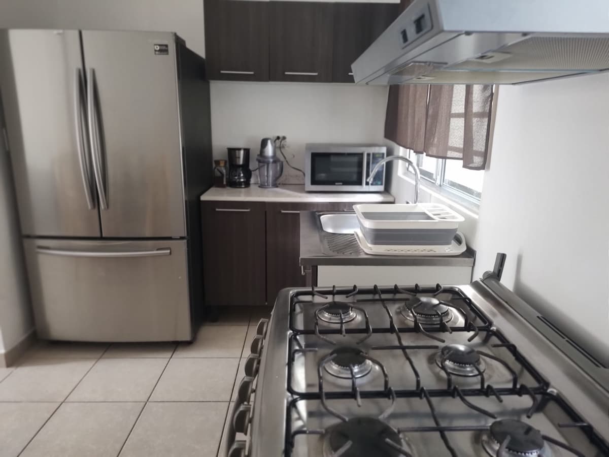 A modern kitchen features stainless steel appliances, including a refrigerator and microwave. A gas stove with multiple burners is positioned prominently, complemented by a stainless steel sink. Natural light enters through a nearby window, enhancing the space's functionality.