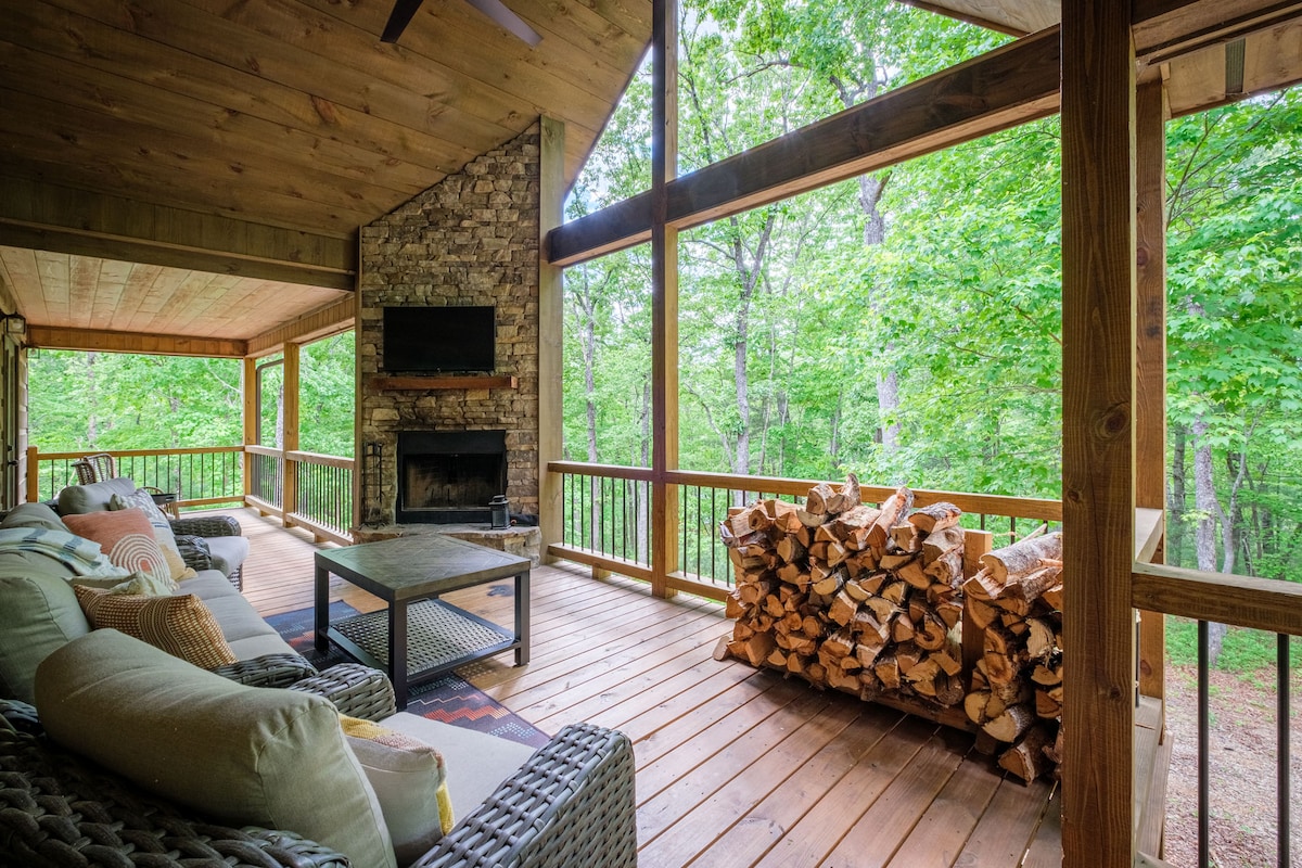 A spacious covered deck features comfortable seating arranged around a coffee table, with a stone fireplace and TV nearby. A stack of firewood sits neatly to one side, while large windows offer views of the surrounding lush greenery.