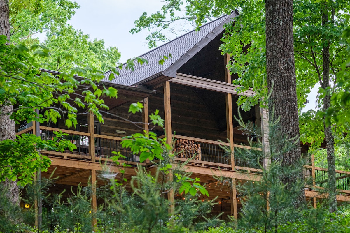 A modern cabin is nestled among lush trees, featuring a covered porch with wooden railings. The peaked roof rises above the natural surroundings, with hints of outdoor lighting visible through the greenery. The combination of natural wood and greenery creates a serene outdoor environment.