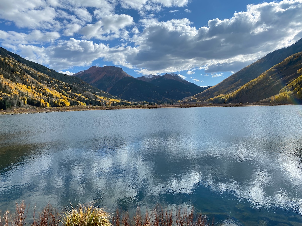 A serene lake reflects the surrounding mountains and sky, framed by colorful autumn foliage on the hillsides. The still water captures the interplay of clouds and sunlight, creating a peaceful natural landscape.