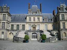 A historical building is presented, showcasing elegant architecture with intricate detail. The entrance features a grand staircase leading up to large glass doors. Flanking the staircase are well-maintained potted plants. The structure is set against a clear blue sky.