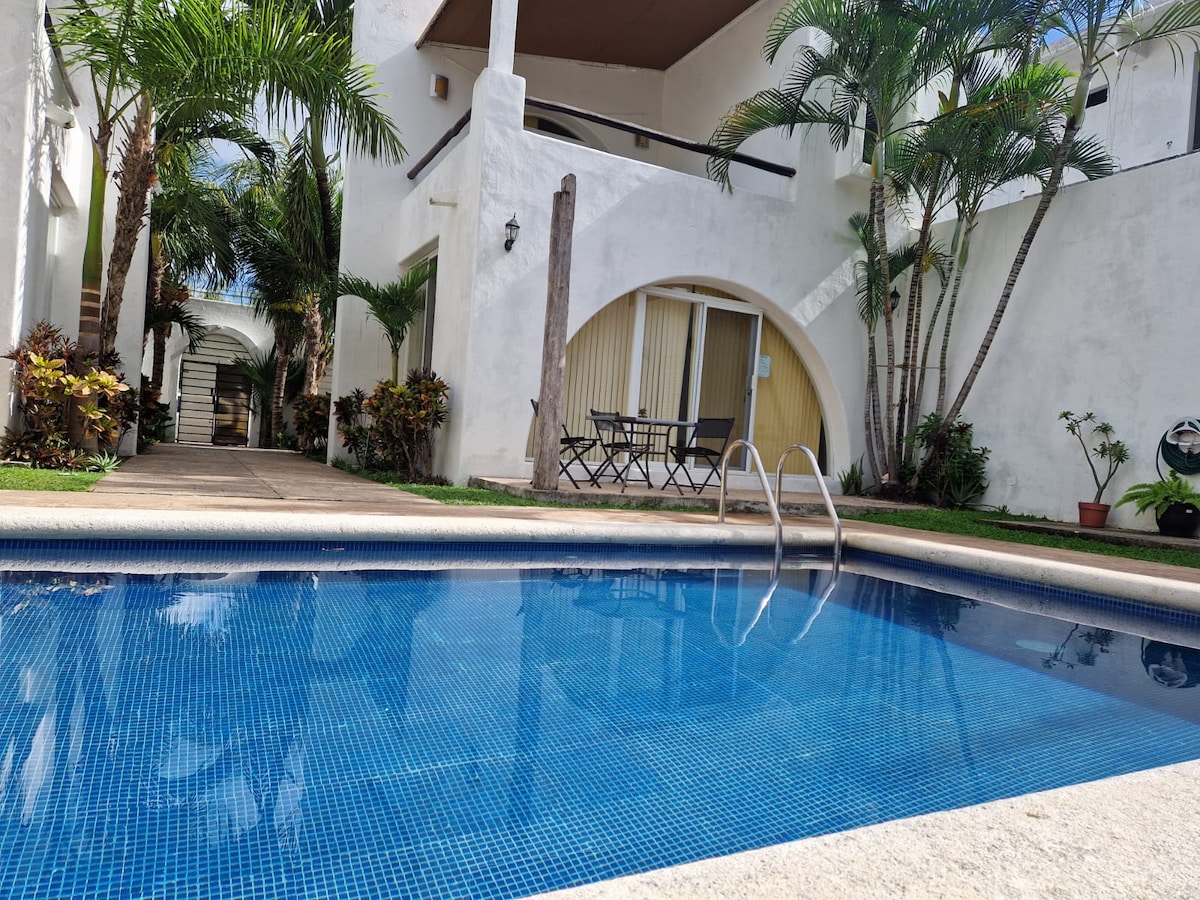 A serene swimming pool is situated in a landscaped area surrounded by palm trees and tropical plants. The pool's clear blue water reflects the sunlight, while an outdoor seating arrangement with a small table and chairs is visible near the entrance of the residence.
