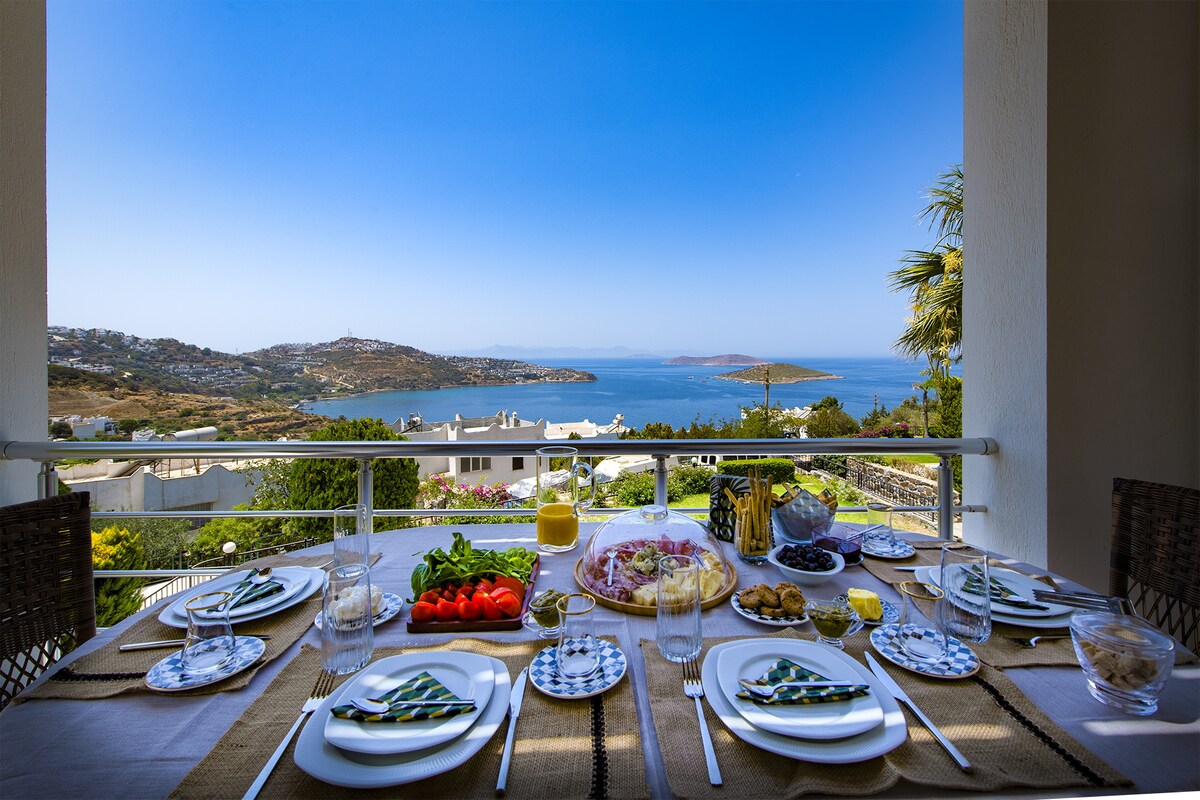 A dining table is set for breakfast with plates, glasses, and cutlery arranged neatly. Fresh tomatoes, olives, and a variety of other foods are displayed, with sea views visible through the railing. The sky is clear, providing a bright backdrop to the outdoor setting.