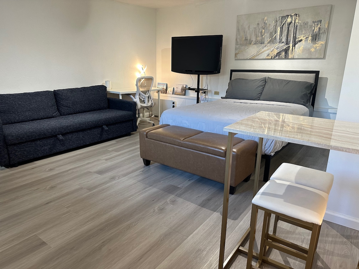 A spacious guest room features a queen bed with a light-colored blanket and a dark sofa bed. A mounted television is positioned on the wall, while a bar table with two stools sits to the right. Natural light illuminates the room and highlights the wood-like flooring.