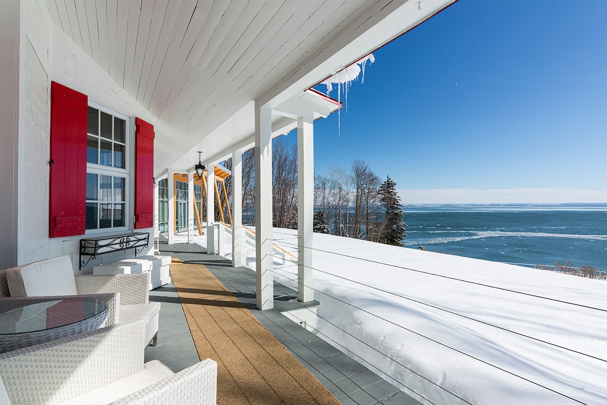 A spacious covered porch features white wicker seating and a round table, providing views of the Saint Lawrence River. Snow blankets the ground, while the blue sky and distant trees create a serene backdrop. Icicles hang from the eaves, reflecting the crisp winter sun.