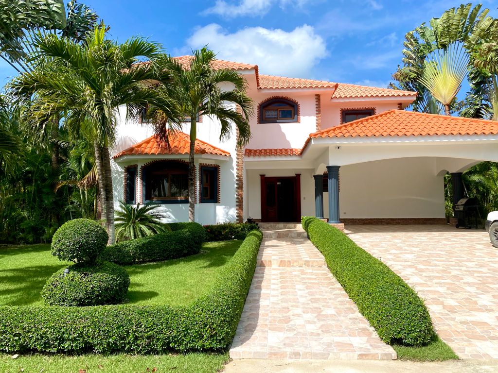 A two-story villa is surrounded by manicured gardens featuring palm trees and neatly trimmed hedges. A stone pathway leads to the entrance, which is accentuated by a covered porch. The orange-tiled roof and white facade create a welcoming appearance.