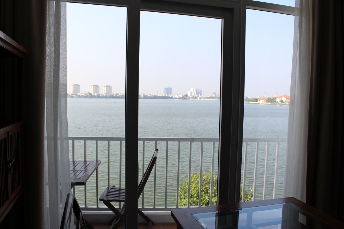 A serene view of a lake is framed by large glass windows, allowing natural light to fill the space. A wooden table and a folding chair are positioned nearby, creating a perfect spot to enjoy the scenic surroundings.