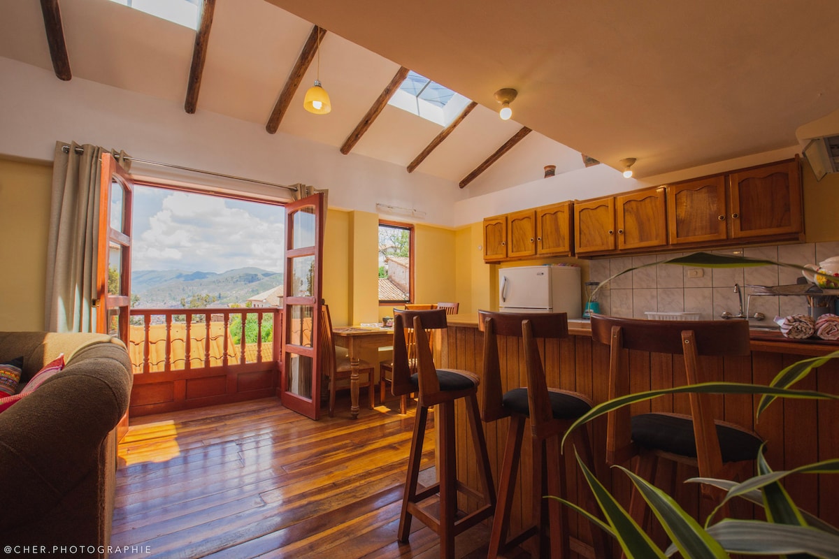 A bright and airy living space features wooden beams and natural light streaming through skylights. An open kitchen showcases wooden cabinetry and appliances, while a dining area is visible. A view of the outdoors is framed by large open doors leading to a balcony, enhancing the inviting ambiance.