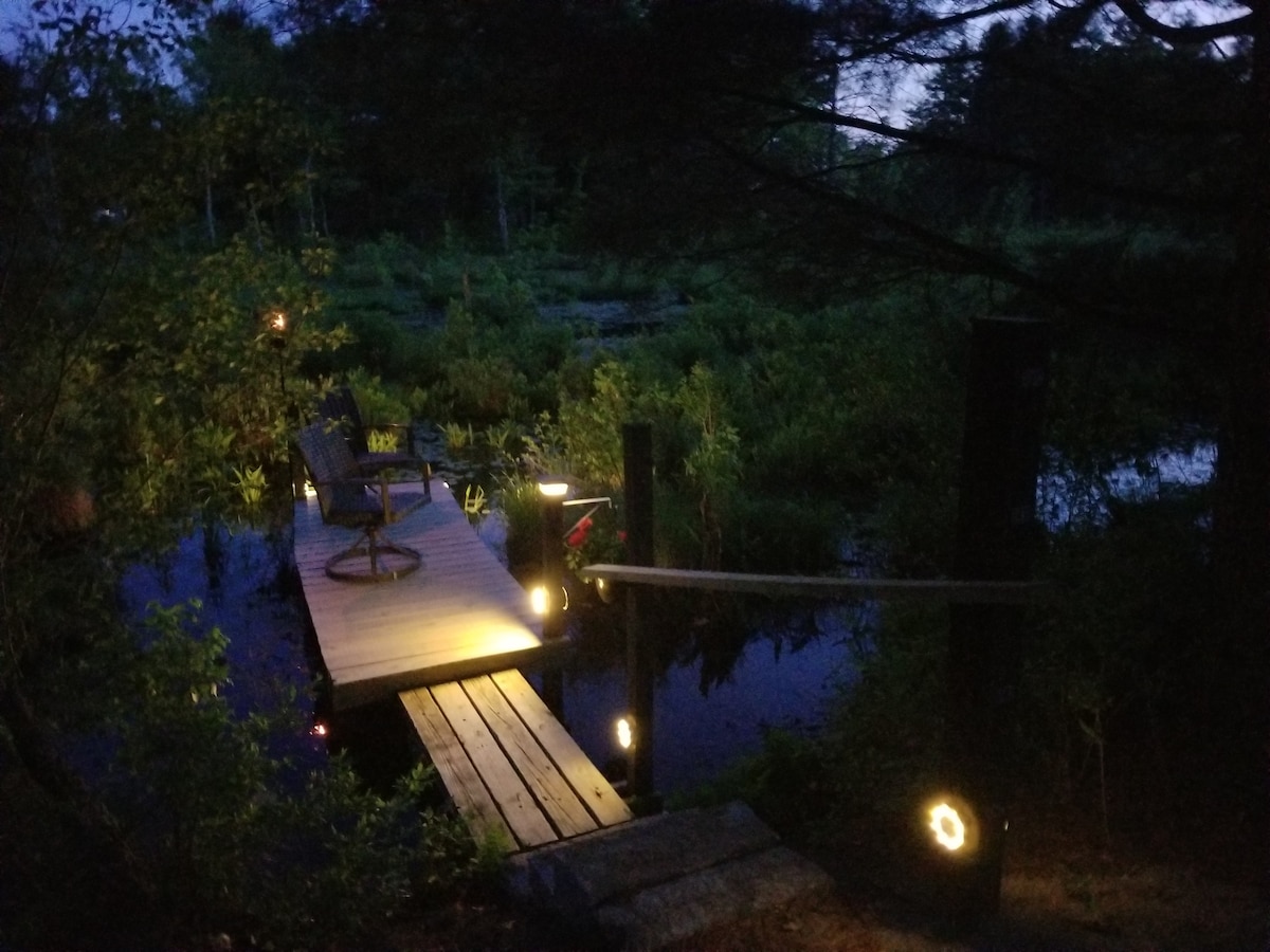 A serene dock extends over tranquil waters, surrounded by lush greenery. Soft lighting illuminates the path and seating area, creating a peaceful ambiance for evening relaxation. The setting invites reflection and appreciation of the natural surroundings.