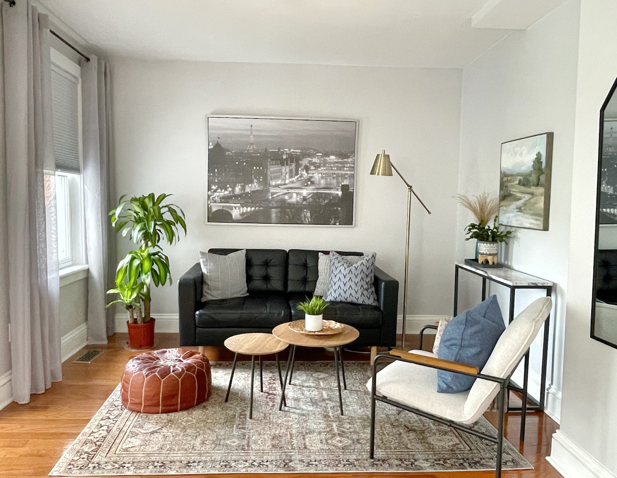 A living area features a modern black sofa paired with two wooden coffee tables and a decorative pouf. A floor lamp stands beside the seating, with a large black-and-white cityscape photo mounted on the wall. Natural light fills the space through the windows, accented by light curtains.