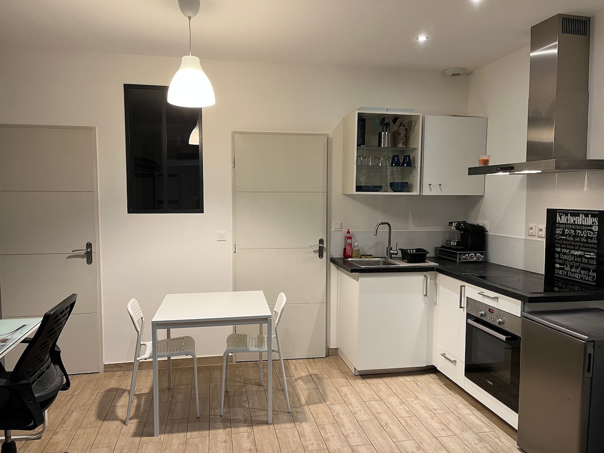 A modern kitchen and dining area is presented with a sleek black countertop and white cabinetry. A small white table with two chairs is positioned in the center, while appliances, including an oven and a sink, are neatly arranged. Natural light enters through a window above the counter.