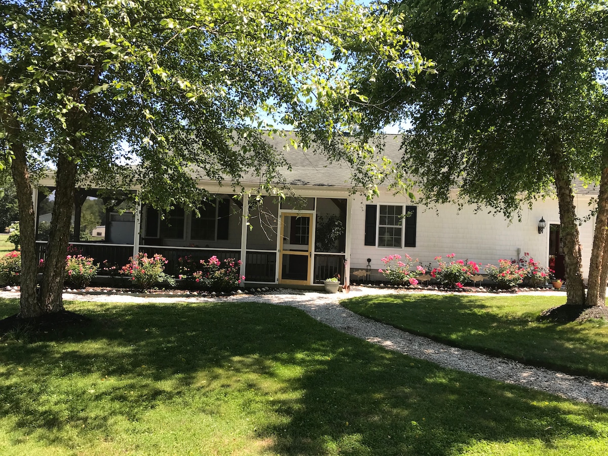 The exterior of the lodge showcases a welcoming entrance with a covered porch framed by flowering bushes. A gravel pathway leads to the front door, surrounded by a lush green lawn and mature trees providing shade.
