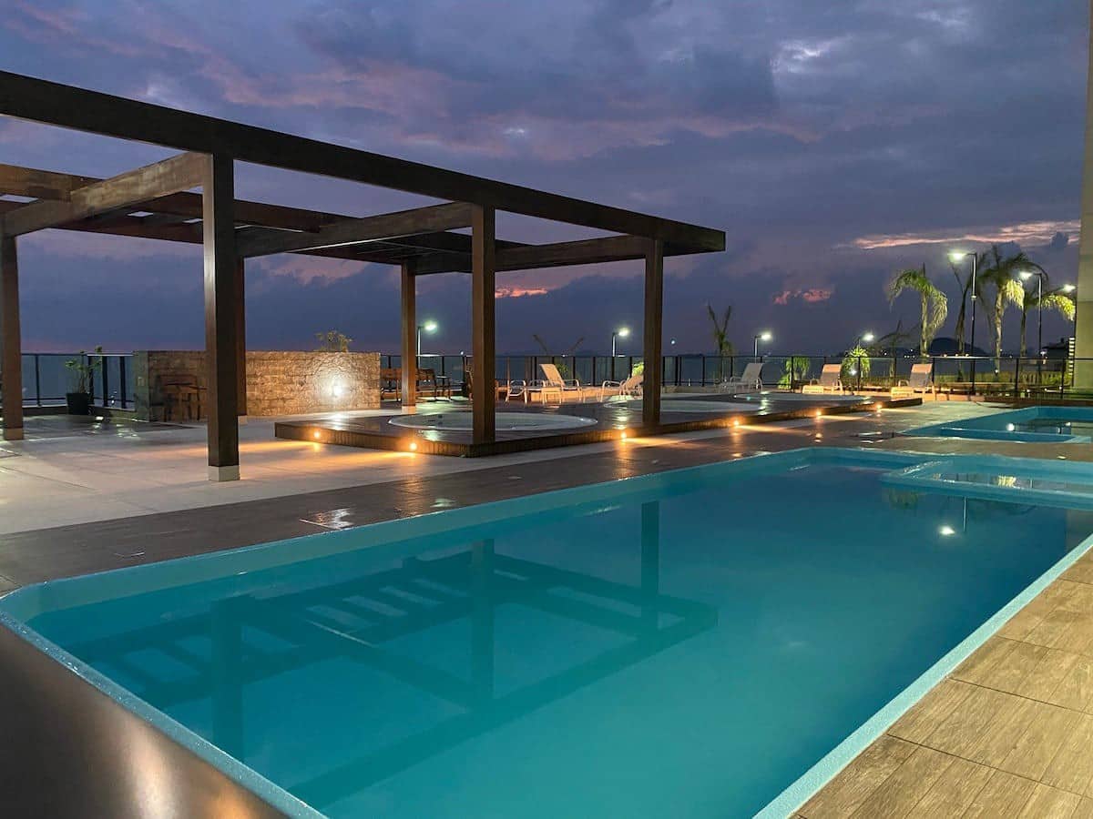 An outdoor pool area features multiple swimming pools, illuminated by soft lighting. Modern lounge chairs and shaded seating areas are arranged around the pools, providing a comfortable setting. The ocean can be seen in the background, with a dusky sky enhancing the serene atmosphere.