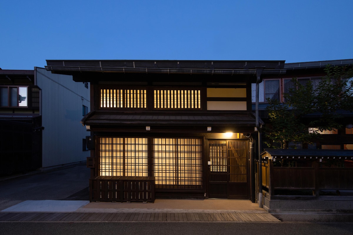 The exterior of the historic townhome features traditional wooden lattice windows, softly illuminated by warm lighting at dusk. The structure is complemented by a simple stone pathway leading to the entrance, creating a welcoming ambiance against the serene evening backdrop.