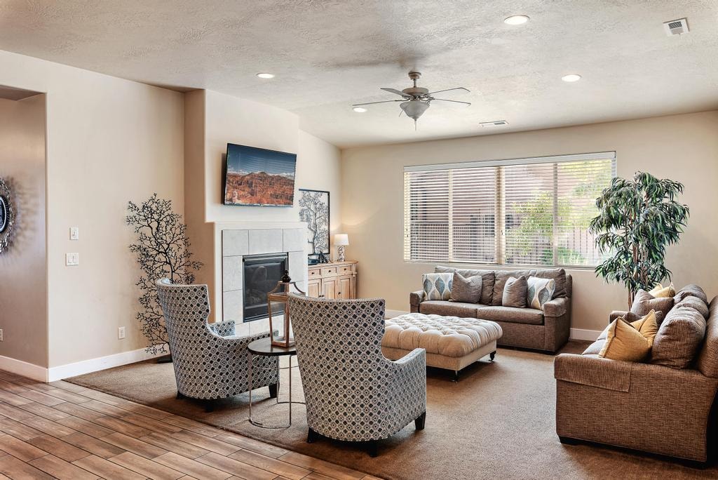 A spacious living area features a comfortable sectional sofa and tufted ottoman, complemented by two patterned armchairs. Natural light fills the room through large windows, with a ceiling fan above and a flat-screen television mounted on the wall. A subtle plant adds a touch of greenery.