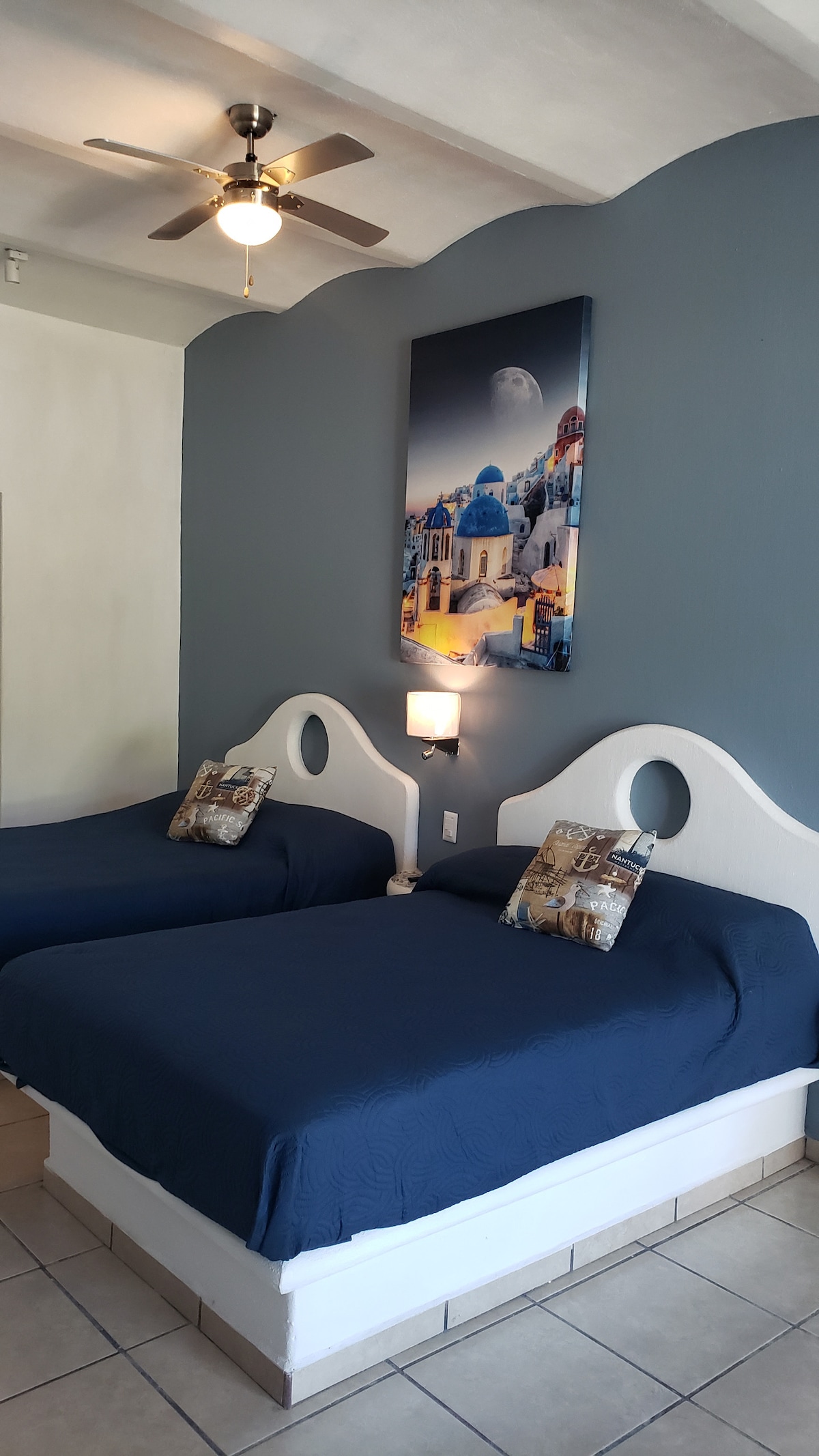 A cozy sleeping area features two spacious beds adorned with navy blue covers and matching decorative pillows. A wall-mounted light fixture provides soft illumination. Above the beds, a serene artwork depicts a quaint village scene, enhancing the restful atmosphere of the room.
